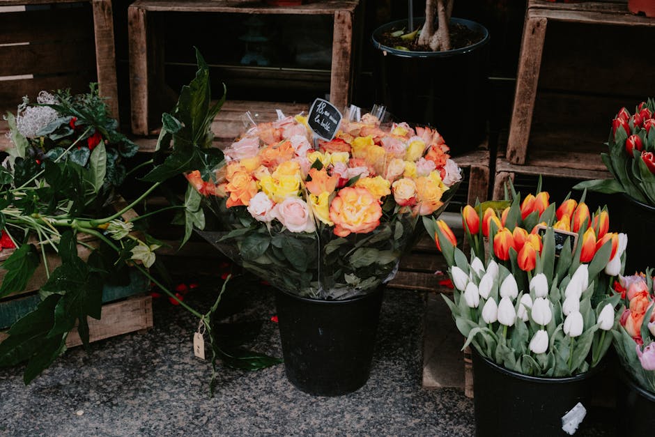 A black ceramic pot filled with a bouquet of vibrant orange, pink, and yellow roses arranged with lush dark green foliage, placed on a patterned surface outdoors. To the right, there is another black 