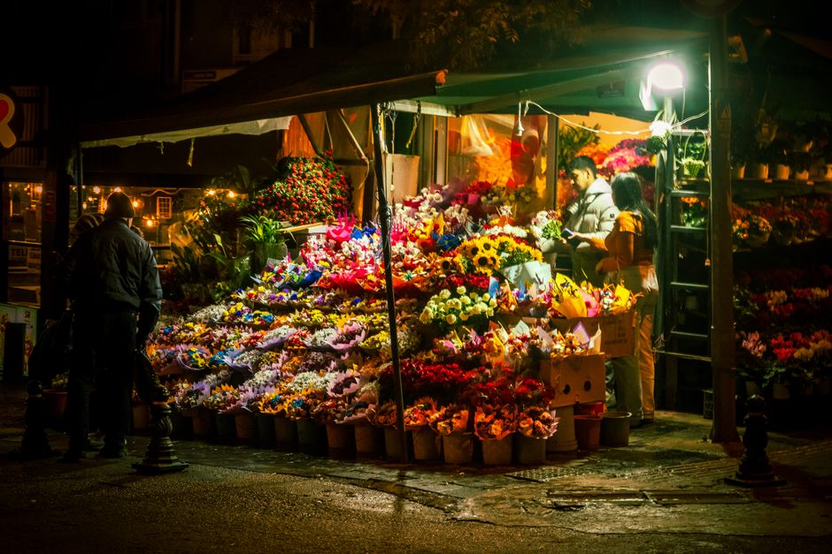 At a flower stall illuminated by warm artificial lighting at night, an array of vibrant, fresh flowers including sunflowers, roses, lilies, and daisies are arranged in buckets and bouquets on multiple