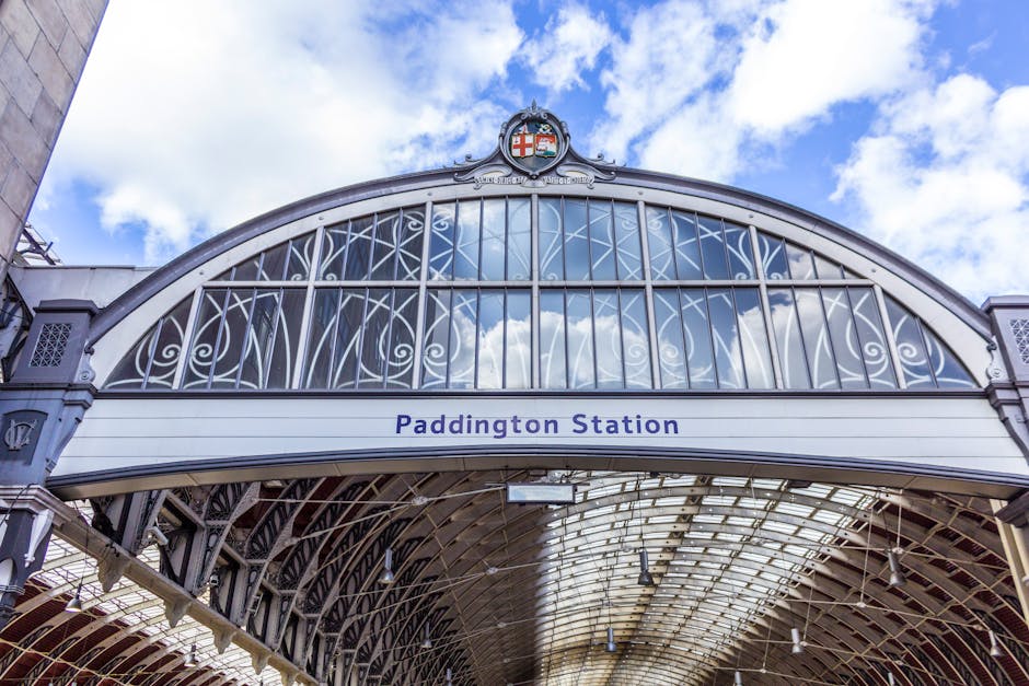 A close-up of a floral arrangement featuring a bouquet of fresh flowers held by a person in front of Paddington Station's iconic ornate arched glass roof and decorative metalwork. The bouquet includes