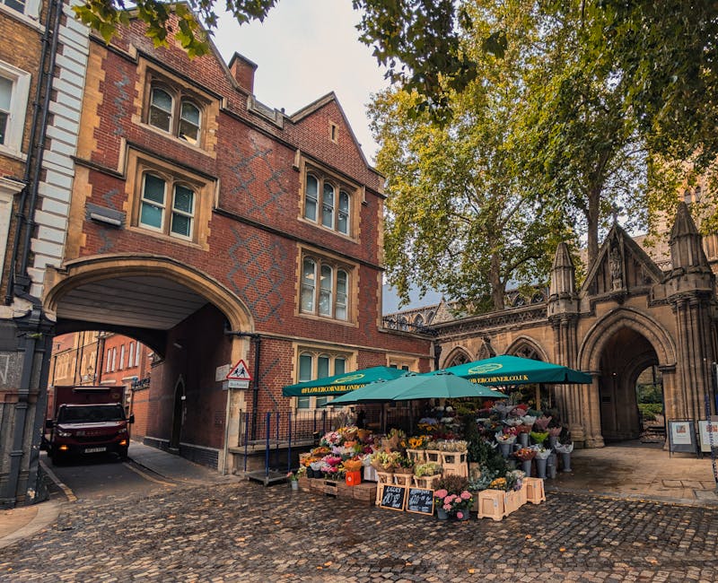 A flower vendor's outdoor stall located on a cobblestone street near a historic brick building with arched walkways, featuring a green umbrella emblazoned with the company’s website. The stall displ