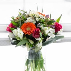 A sophisticated bouquet, meticulously crafted by local Paddington florists, is captured in the embrace of soft, diffuse daylight that pours through the windows of Sussex Gardens or nearby cafés. The heart of the arrangement is a chorus of gerbera daisies in radiant orange and creamy beige, their layered petals imbued with both energy and sophistication. Embracing them, magenta roses lend structure and decadent allure, each intricate spiral a testament to calm devotion. Clusters of white alstroemeria and fine spray roses are woven with expert care, bringing notes of purity and gentle radiance to the arrangement. Gentle hints of blush pink roses peek out, introducing a playful yet refined nuance. Oversized, glossy aspidistra leaves curve with purposeful grace, anchoring the design and recalling Paddington's green, riverside appeal. Sprigs of lighter foliage fill in between, their fresh texture crafting a sense of lush abundance. Encased in a tall, clear glass vase, verdant stems are elegantly displayed, echoing the transparency and freshness that define the area's floristry. Backlit by a serene white glow suggestive of early light over Little Venice, each hue and petal stands out, creating an arrangement that feels at once celebratory and intimate, perfect for gatherings, milestones, or simple gestures of gratitude towards neighbours in the community.